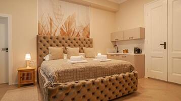 A bedroom with a large bed, a bedside table with a lamp, a wall-mounted artwork, a kitchenette, and a door.