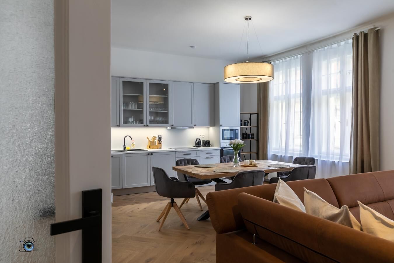 Una cocina moderna con armarios grises, un comedor con mesa y sillas de madera, y un sofá marrón en primer plano.