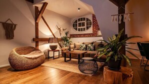 Una sala de estar acogedora con suelo de madera, un sillón orejero tejido, una mesita de centro redonda y una planta grande en maceta.