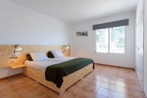 A bedroom with a wooden headboard, a bed with a green blanket, two bedside lamps, and a window with a view of buildings. - Neighbours Apartments (Calvia)