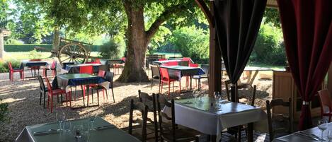 Un espacio de comedor al aire libre con mesas y sillas bajo un techo cubierto, rodeado de árboles y vegetación.