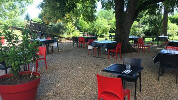 Un área de comedor al aire libre con mesas y sillas dispuestas sobre grava, rodeada de árboles.