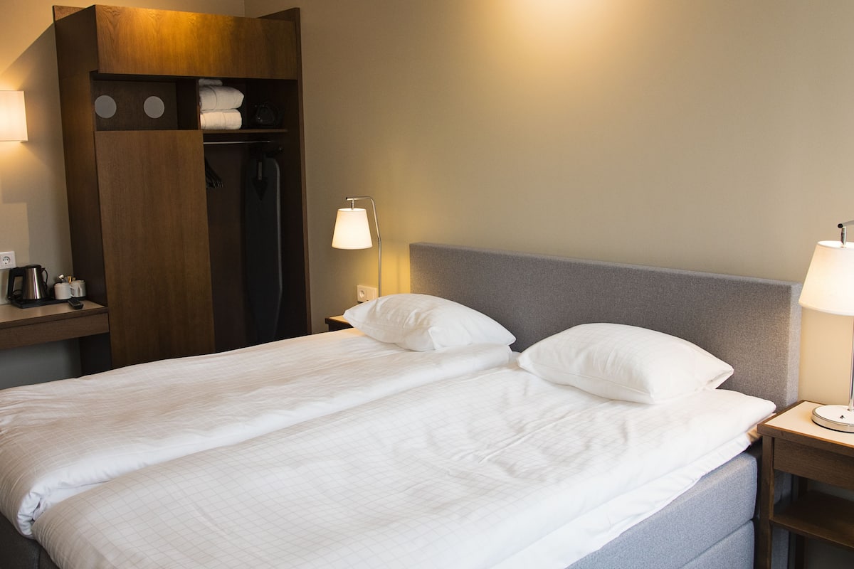 A neatly made bed with two pillows, a bedside table with a lamp, and a wooden wardrobe with a mirror and a hanging coat.