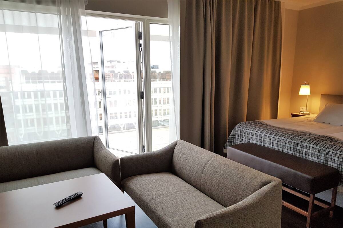 A hotel room with a sofa, a small table, a bed with a checkered blanket, a nightstand with a lamp, and a view of buildings through the window.