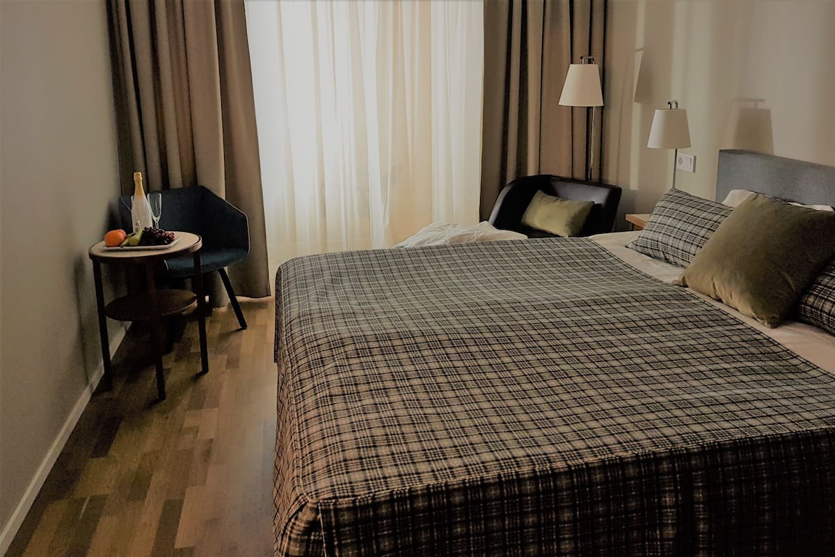A hotel room with a large bed, a small table with a bottle and fruit, a chair, and a window with curtains.