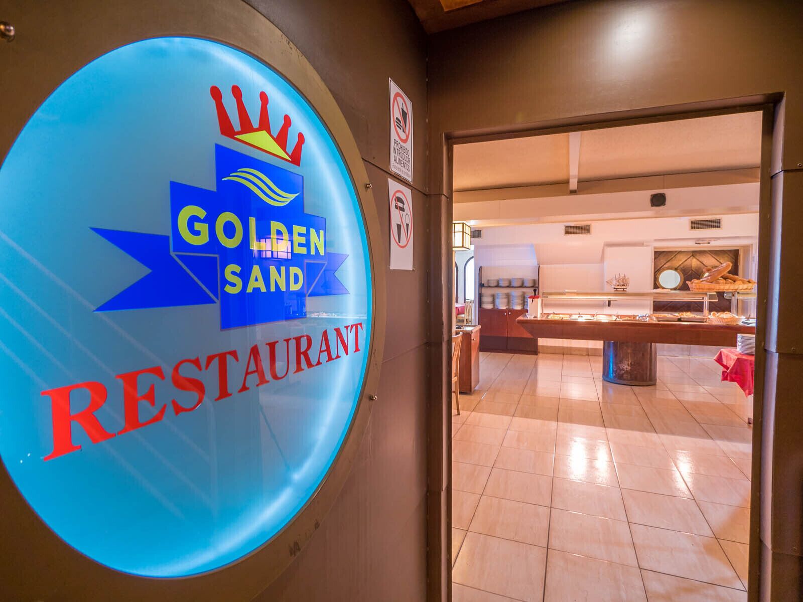 Une enseigne de restaurant avec les mots « GOLDEN SAND » et « RESTAURANT », un panneau indiquant qu’il est interdit de fumer, et un aperçu de l’intérieur, où l’on aperçoit un buffet.