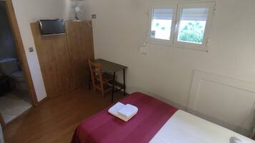 A small room with a bed, a wooden wardrobe, a small table, a chair, a TV on the wardrobe, and a window with a view of greenery.