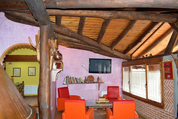 Un interior rústico con vigas de madera, un televisor montado en la pared, una estantería con libros y objetos decorativos, y sillas anaranjadas alrededor de una mesa.