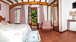 Ein Schlafzimmer mit einem großen Bett, einem hölzernen Kopfteil, einem Nachttisch, einem Sessel, einem kleinen Tisch mit einer Vase und Blick ins Grüne durch eine geöffnete Tür.