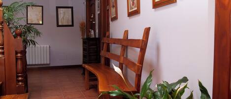 A wooden bench, a potted plant, and framed pictures on a wall in a room with a tiled floor and wooden ceiling beams.