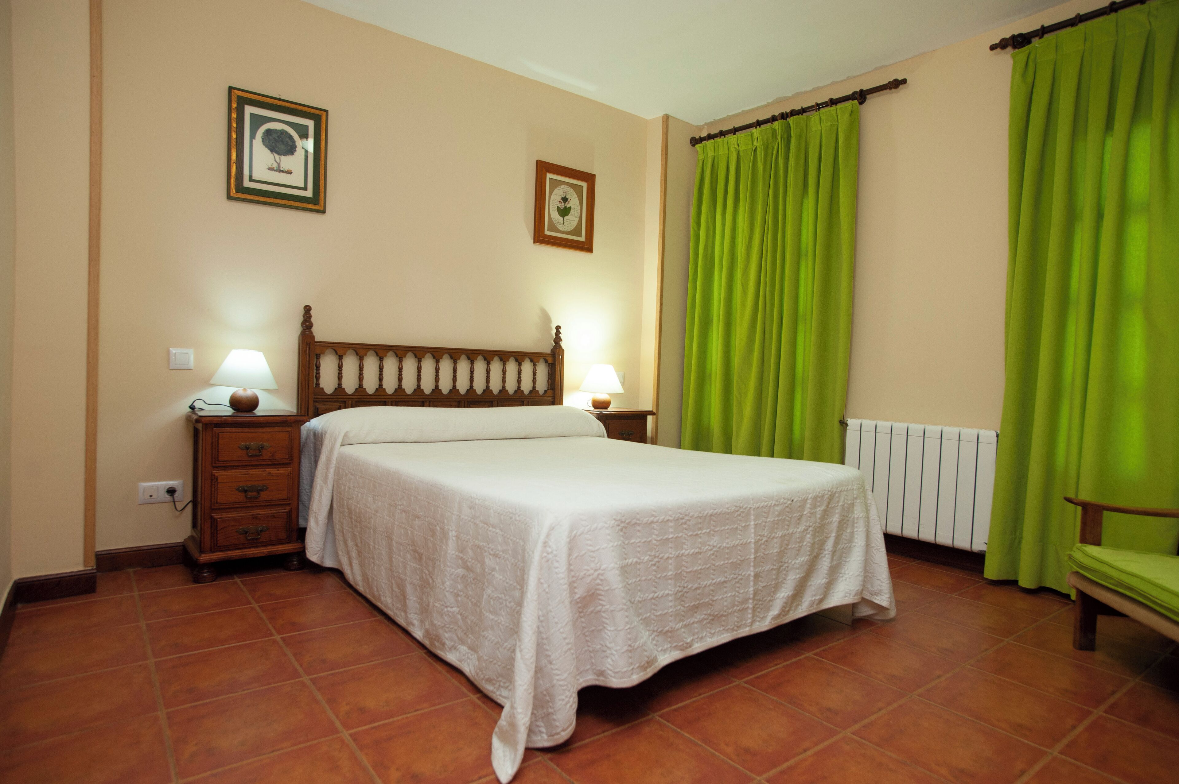 Un dormitorio con una cama, dos mesitas de noche con lámparas, una mesita auxiliar de madera, un radiador y una silla.