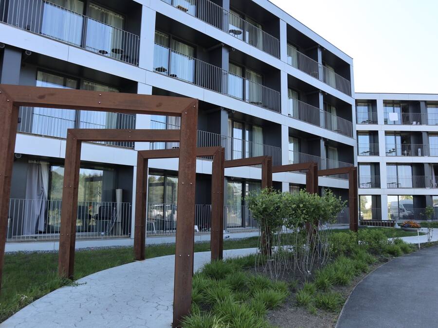 Un edificio moderno de varios pisos con balcones, un paseo pavimentado y un pequeño jardín.