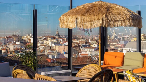 Una terraza en la azotea con mobiliario de mimbre, un parasol de paja y vistas de la ciudad.