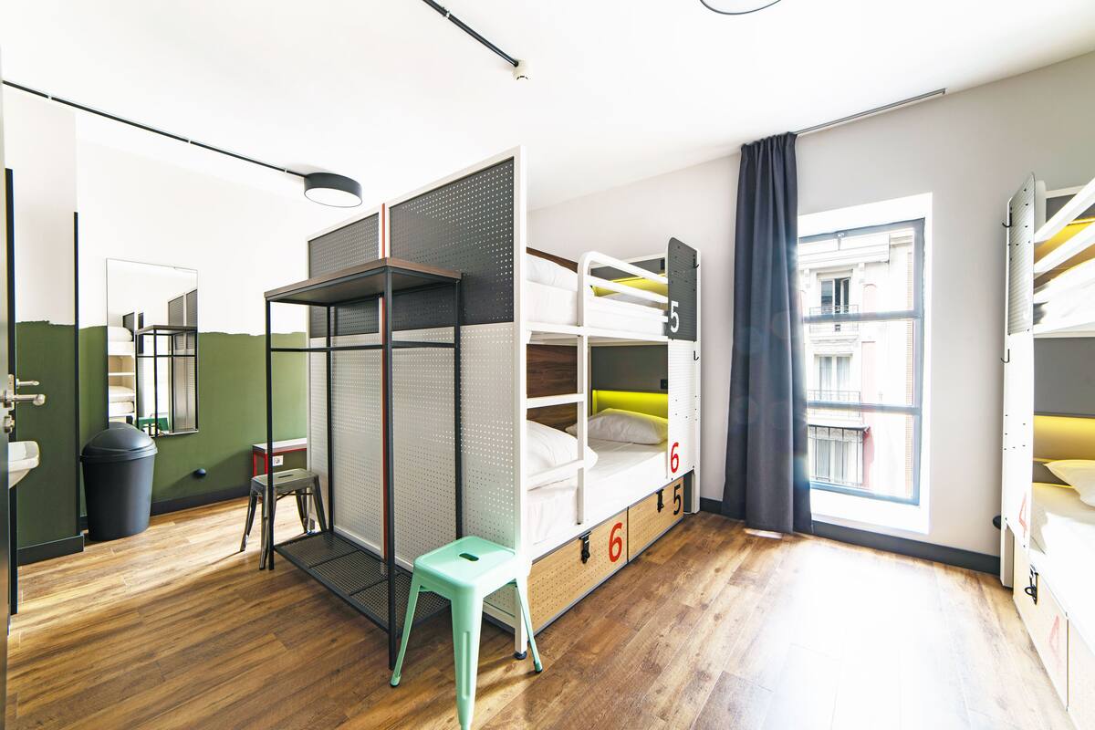 Habitación de hotel compacta con literas, un taburete pequeño, un bote de basura y una ventana con vistas a edificios.