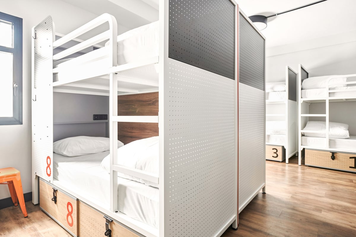 Habitación con literas, suelo de madera, un taburete pequeño naranja y una puerta con el número 8.