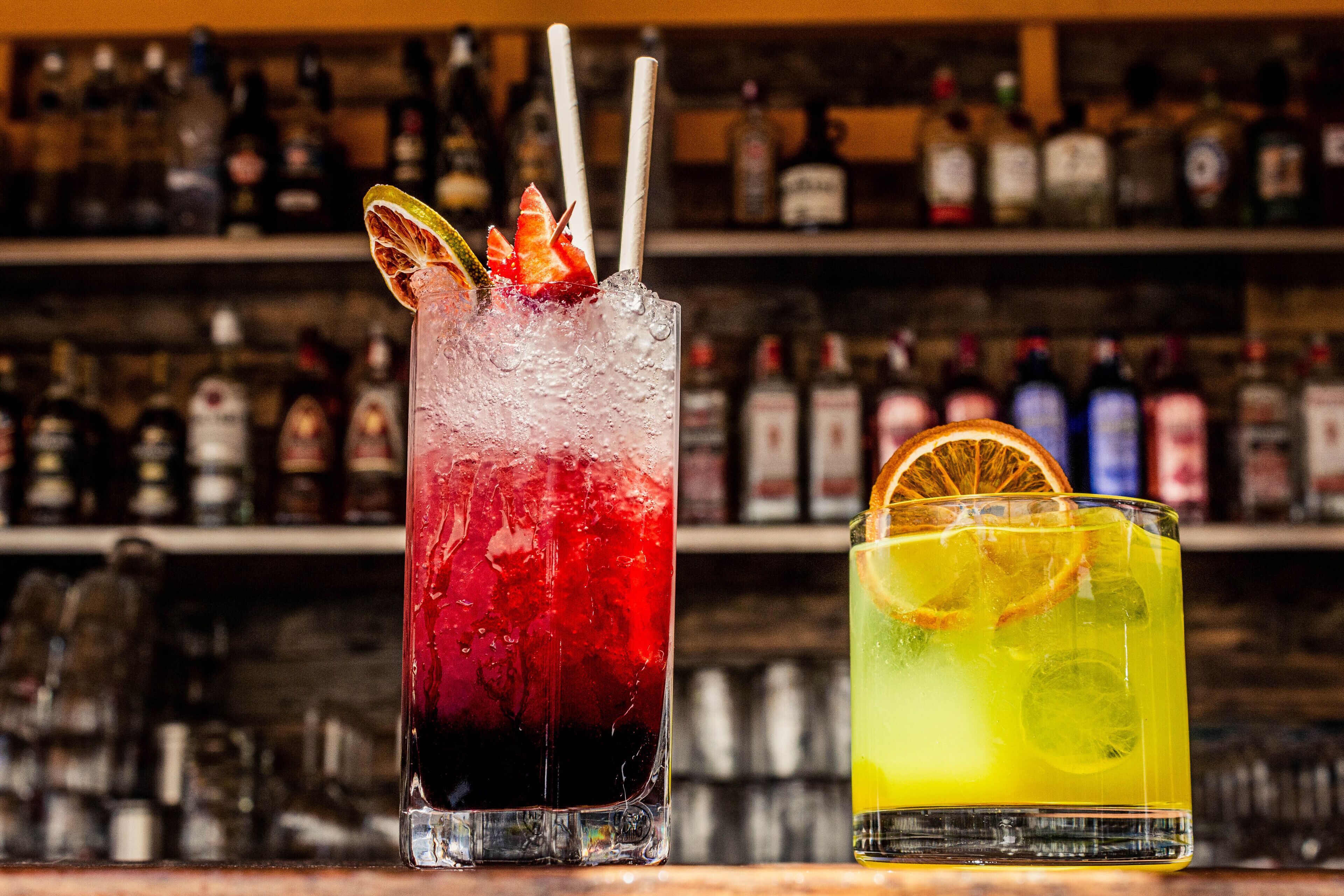 Two cocktails with straws and garnishes, a bar with shelves of bottles in the background.