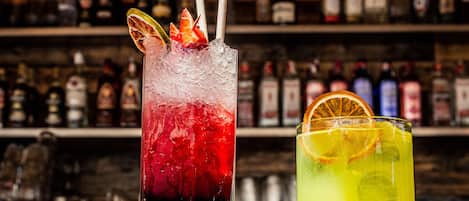 Two cocktails with straws and garnishes, a bar with shelves of bottles in the background.