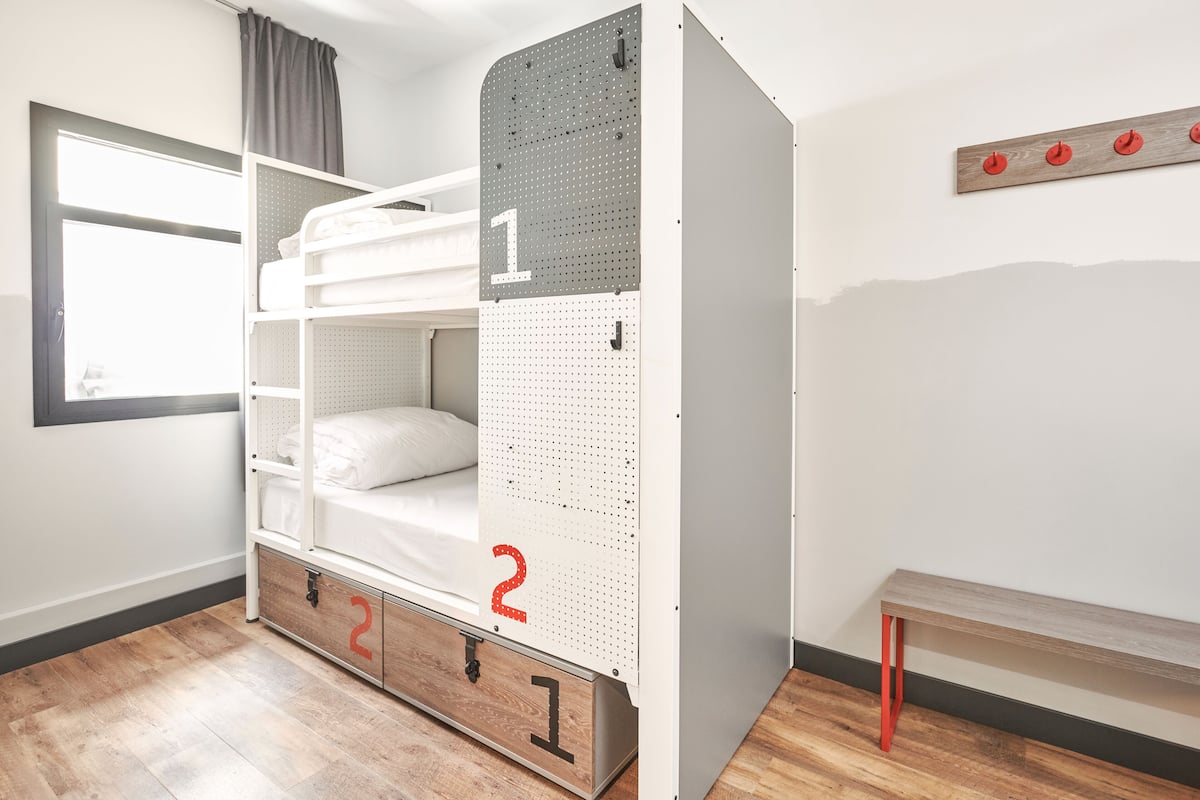 Una cama litera con un panel perforado, un banco y una ventana con cortinas.