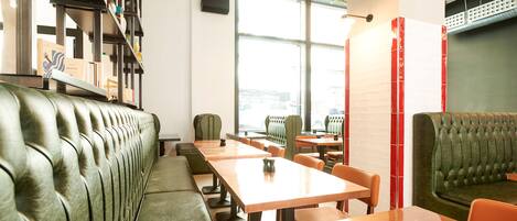 A modern restaurant with green upholstered booths, wooden tables, and a large window offering a view of the city.
