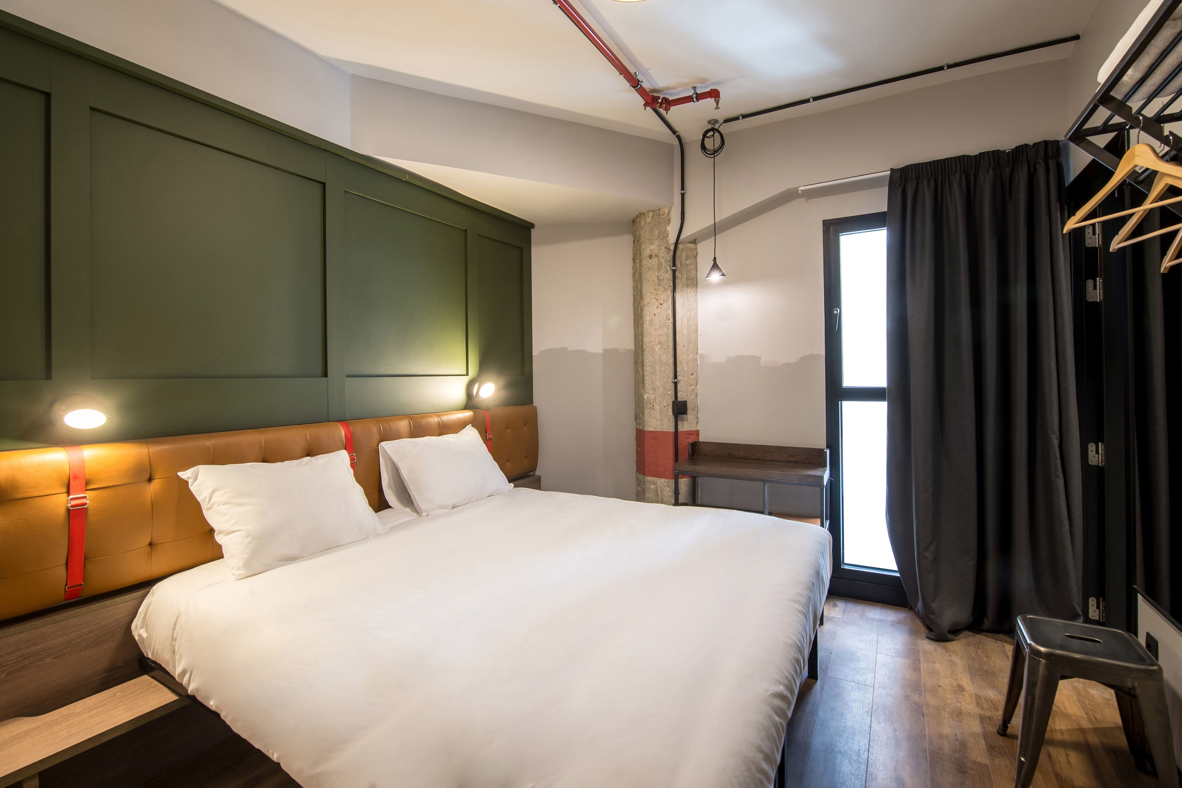 A hotel room with a large bed, a wooden bench, a hanging light fixture, and a window with curtains.