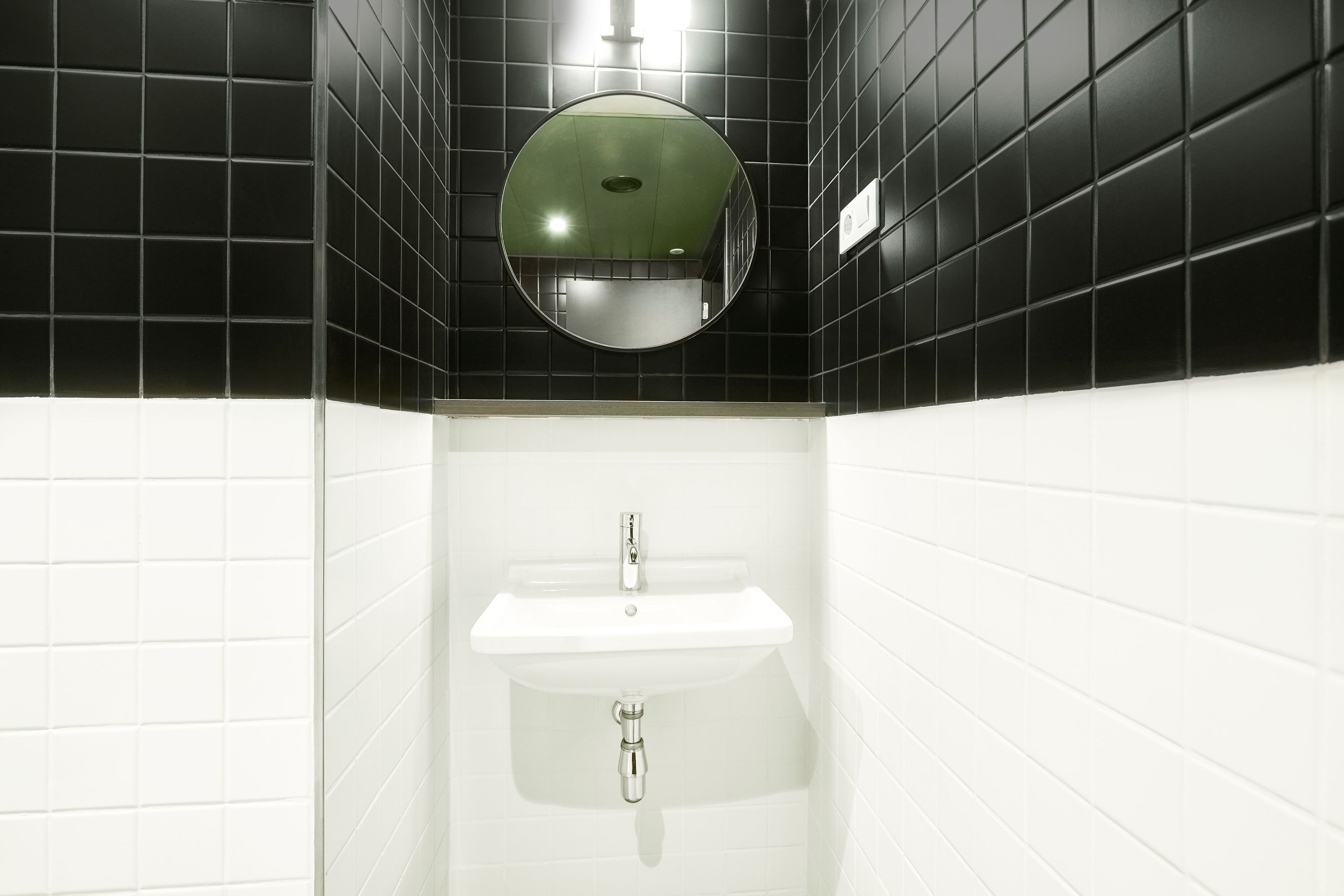 Un baño con paredes de azulejos blanco y negro, un espejo redondo y un lavabo blanco con grifo.