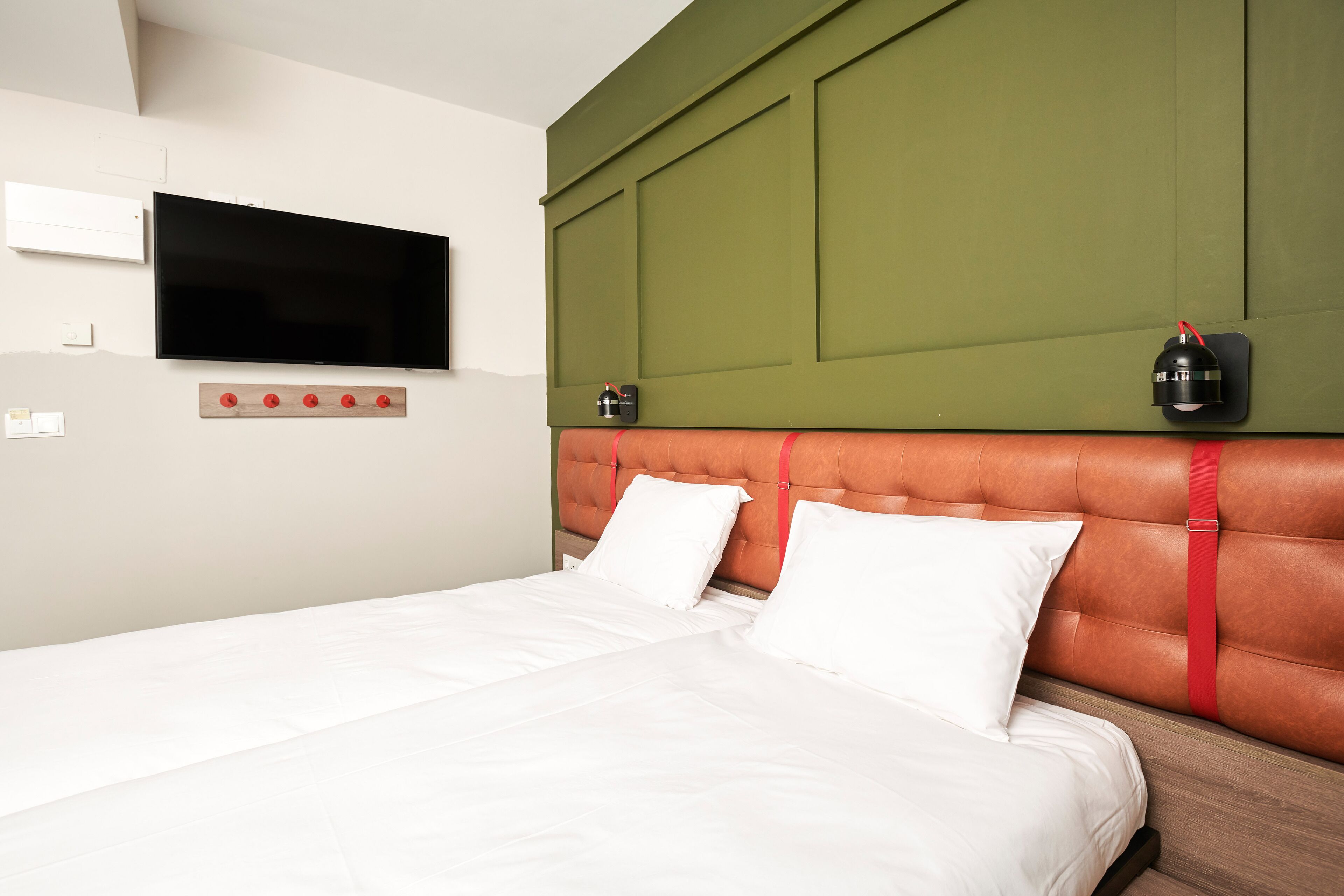 A neatly made bed with white linens, a mounted flat-screen TV, and two wall-mounted lamps.