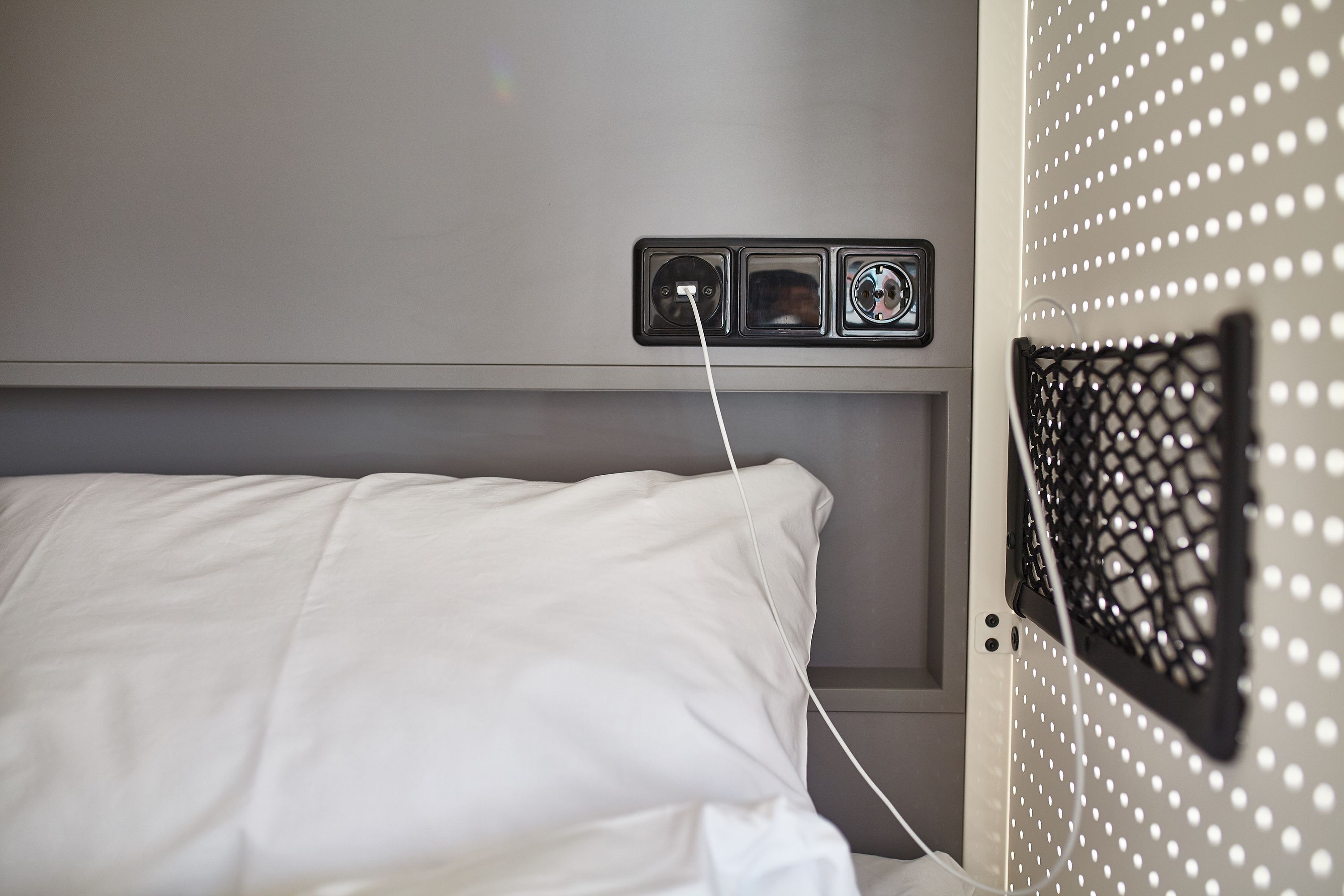 Un lit avec des draps blancs, une prise de courant avec une fiche, et un écran en treillis fixé au mur.