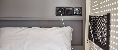 Un lit avec des draps blancs, une prise de courant avec une fiche, et un écran en treillis fixé au mur.