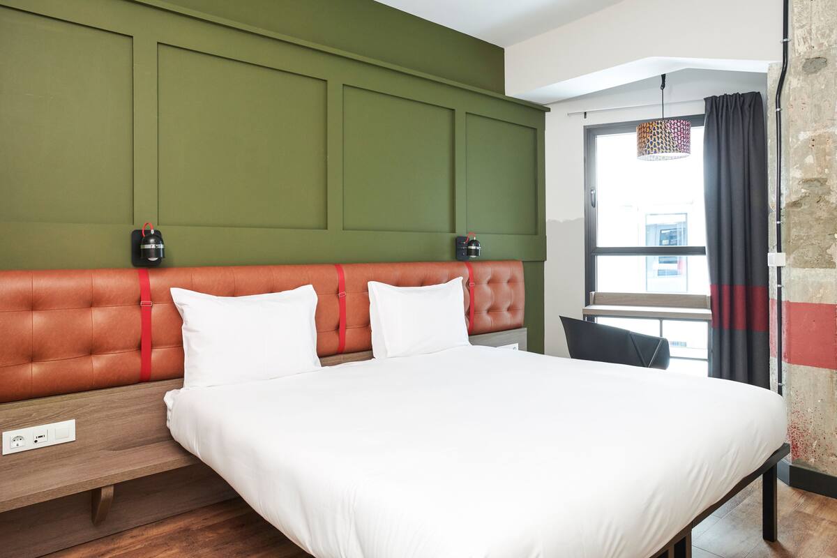 Una habitación de hotel moderna con una cama grande, paredes de acento verde y vistas a la ciudad a través de una ventana.