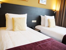 Quarto de hotel com duas camas, cada uma com roupa de cama branca e almofada estampada. Há uma mesa de cabeceira com um telefone e um controle remoto.