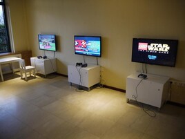 Uma sala de jogos com três consoles exibindo diferentes jogos, uma mesa branca e uma janela.