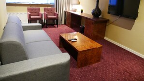 Une chambre d’hôtel avec un canapé gris, deux fauteuils, une table basse en bois, un téléviseur et une lampe.