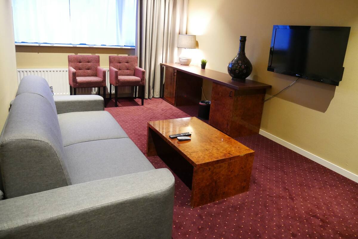 Habitación de hotel con sofá gris, dos sillones, una mesa de centro de madera, televisión y lámpara.