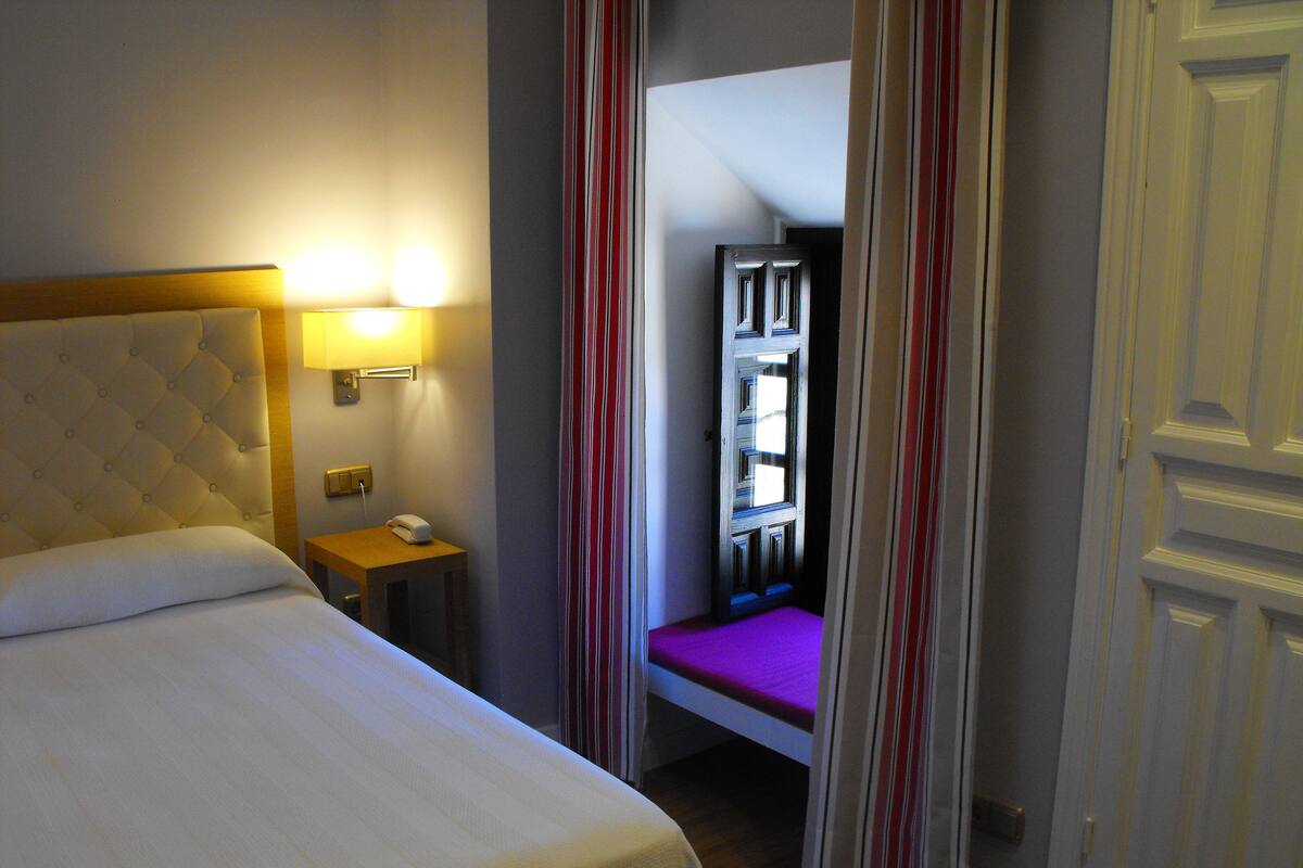 A hotel room with a bed, bedside table, lamp, and a view through a door.