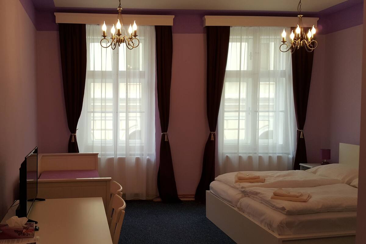 Un dormitorio con cama, escritorio y silla. Cuenta con dos ventanas con cortinas y candelabros.