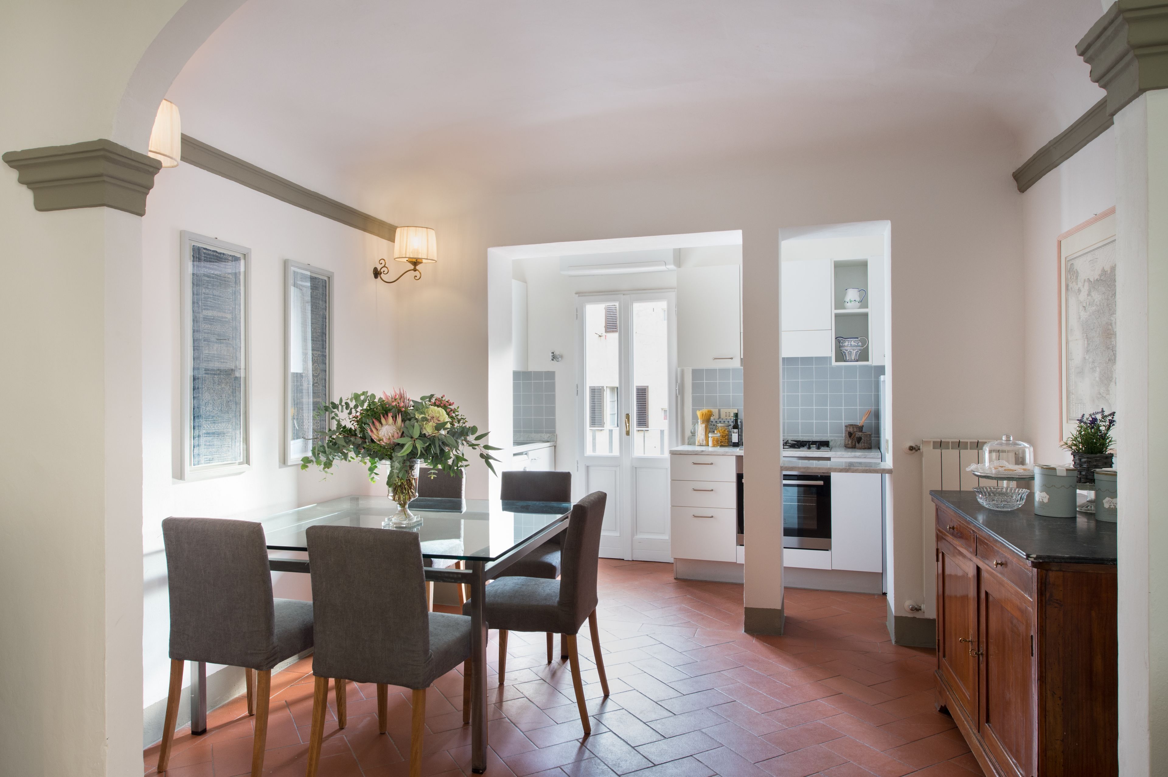 A modern kitchen with a dining area, featuring a glass table, grey chairs, and a vase with flowers.