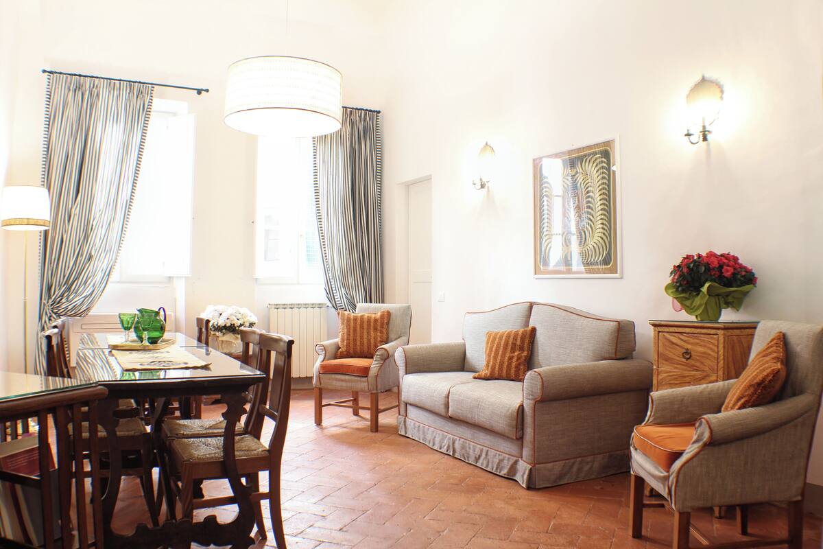 A dining area with a table, chairs, and a chandelier. There are two sofas and a side table with a vase of flowers.