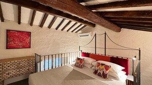 Un dormitorio con techo de madera, cama con ropa de cama blanca y almohadas decorativas, cabecera roja y un cuadro abstracto rojo en la pared.