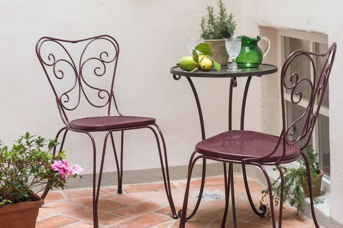 A small patio area with two metal chairs, a table with a potted plant, a green watering can, and a wall-mounted lantern.