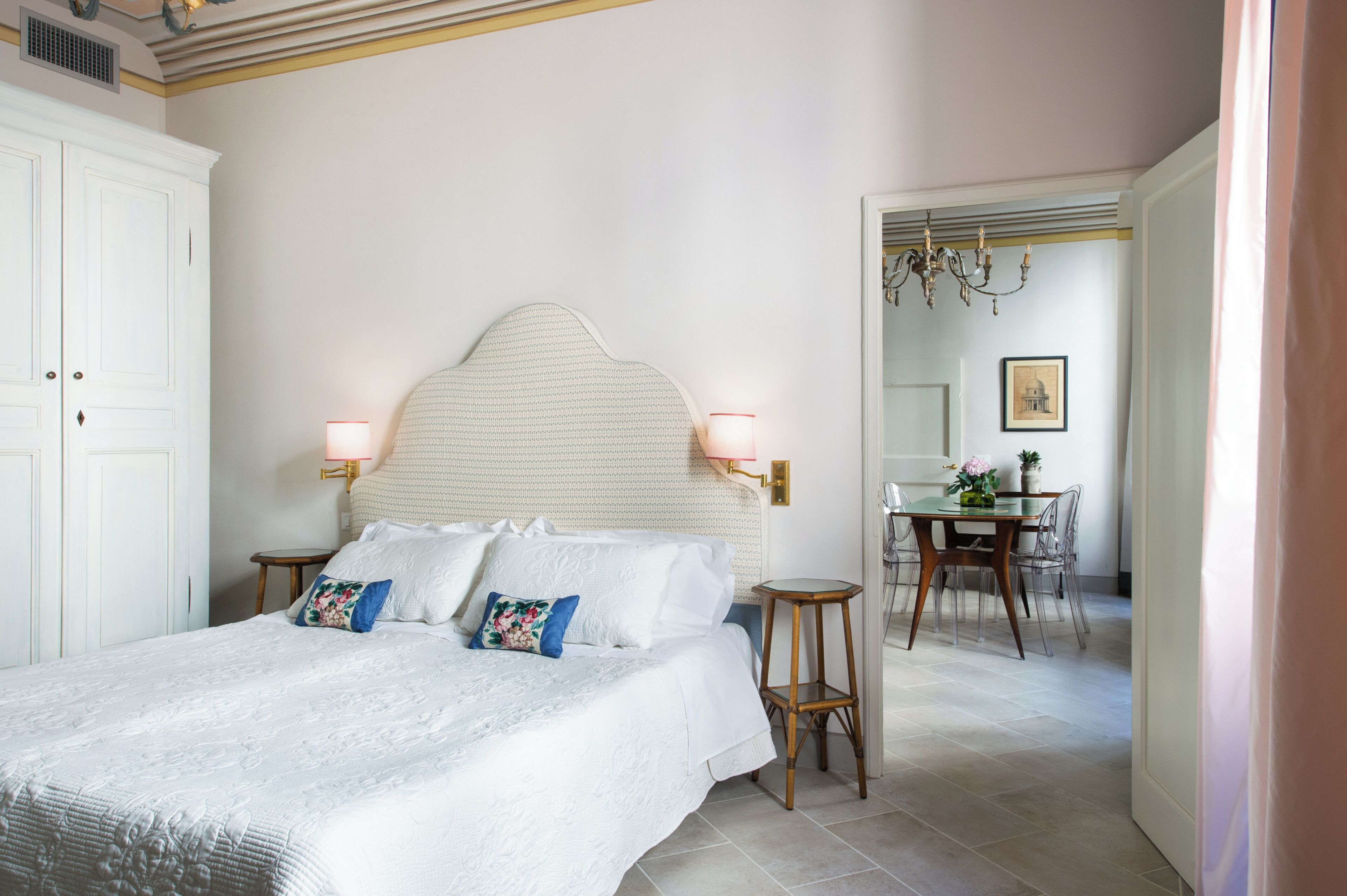 A bedroom with a large bed, two bedside tables, a chandelier, and a dining area with a table and chairs.