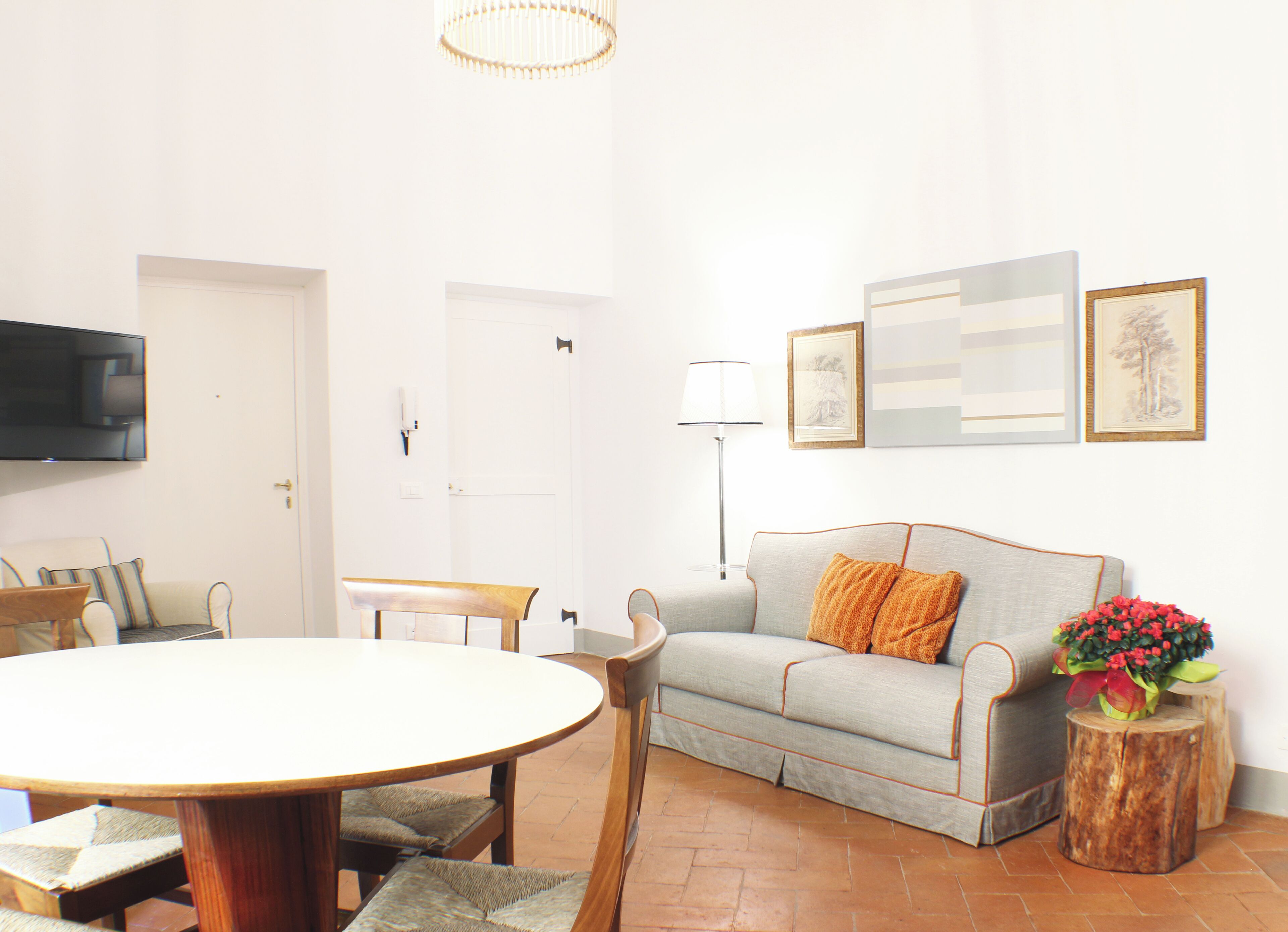 A living room with a round wooden table, a grey sofa with orange cushions, a wooden coffee table, a TV mounted on the wall, and a floor lamp.