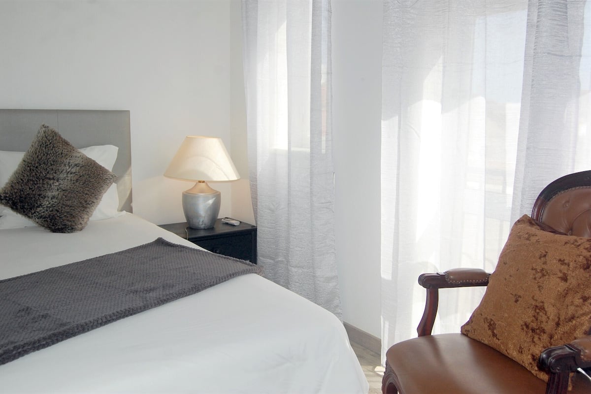 A neatly made bed with a grey throw, a lamp on a bedside table, and a chair with a patterned cushion.