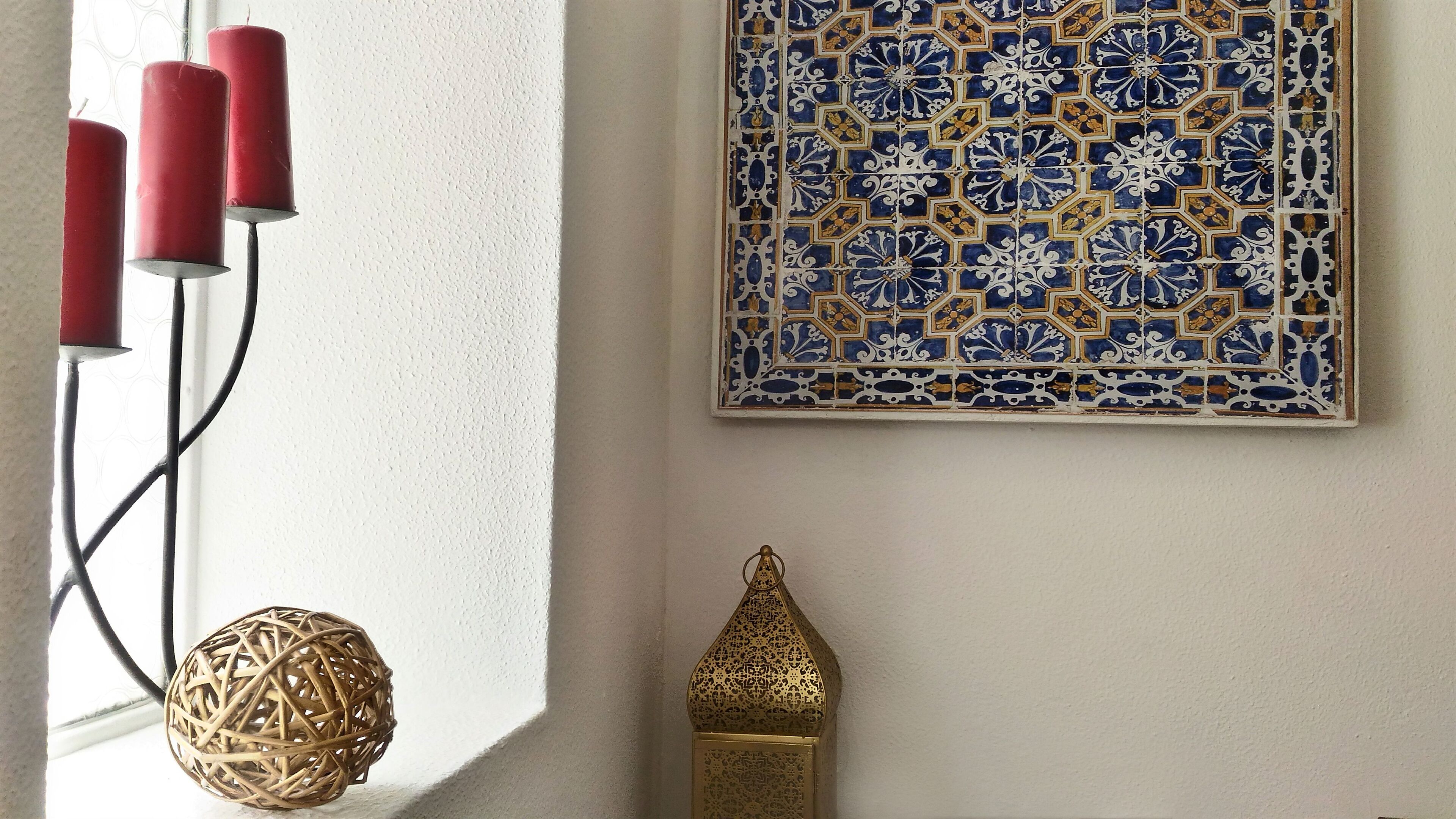 Una pared con azulejos de diseño azul y dorado, un candelabro rojo, un objeto esférico tejido y un adorno dorado.