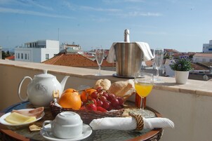 A breakfast setup with a teapot, cups, a basket of fruits, and a bottle of champagne on a balcony overlooking a cityscape.