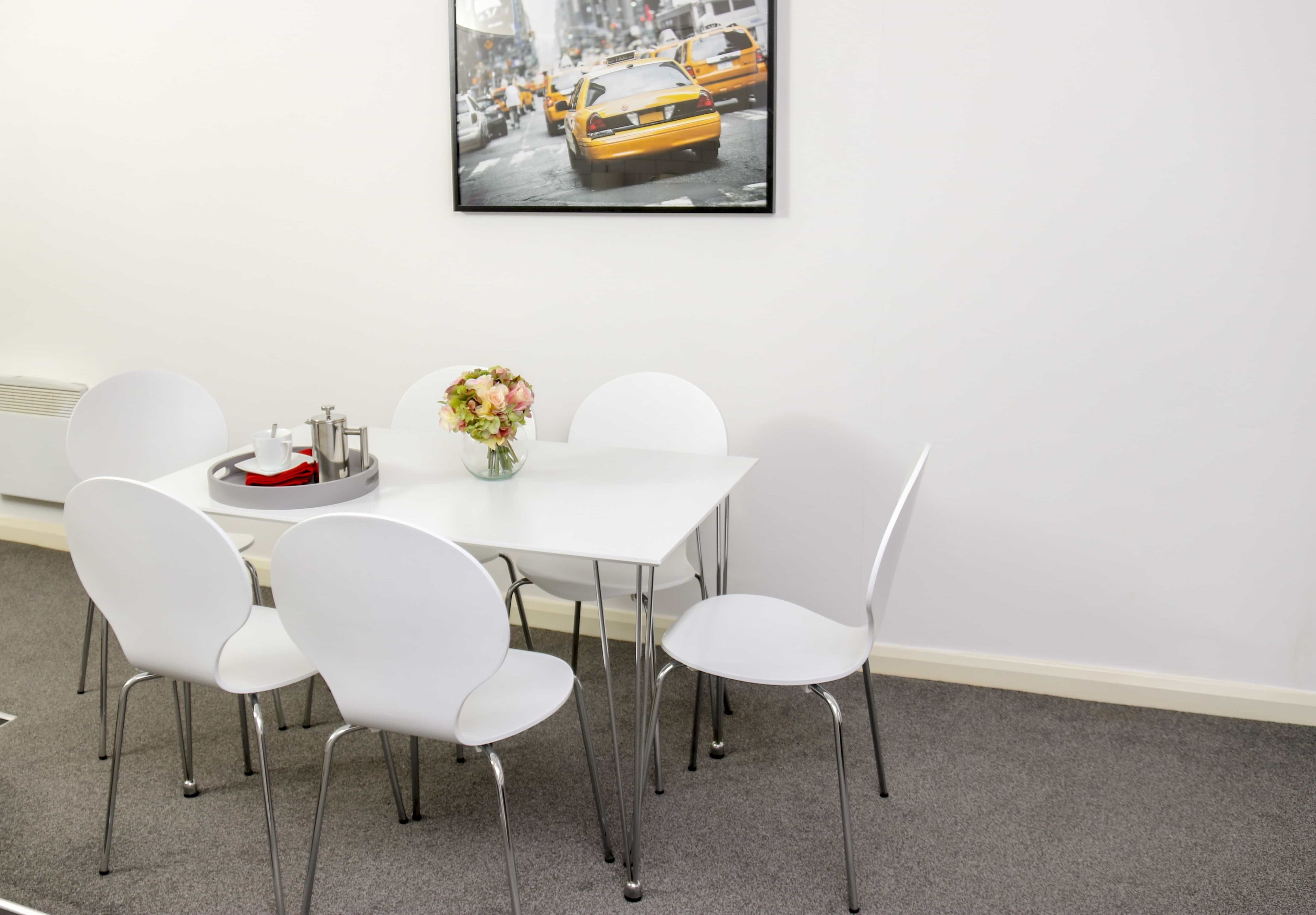A white dining table with four white chairs, a vase with flowers, and a framed picture on the wall.