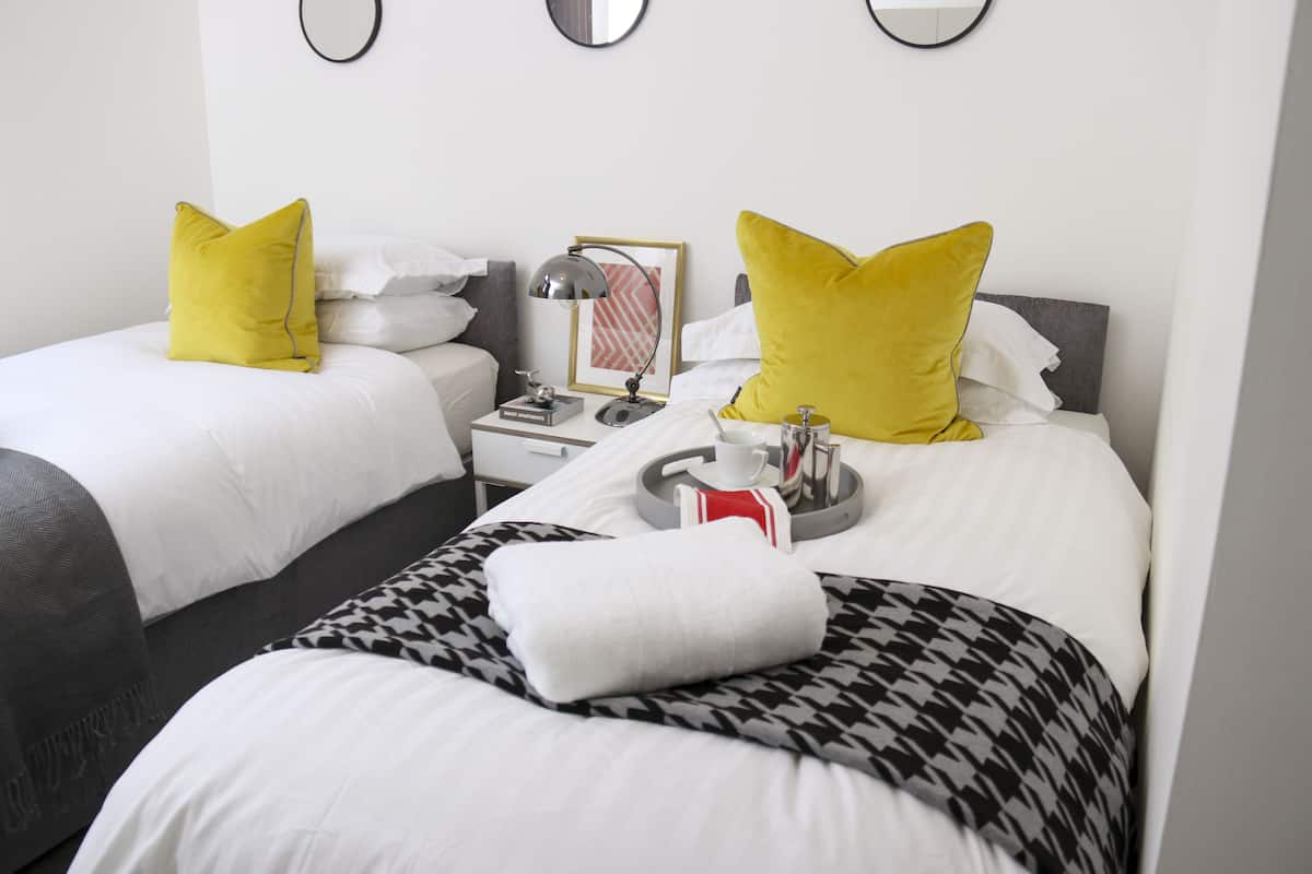 A hotel room with two beds, each with a white and black houndstooth bedspread, and yellow pillows. There is a tray with a cup, a bottle, and a glass on the bed.