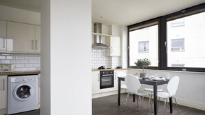 Une cuisine moderne avec des meubles blancs, un four encastré et un lave-linge. On y trouve un coin repas avec une table et des chaises, ainsi qu’une fenêtre donnant sur l’extérieur.