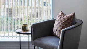 Une pièce moderne avec une grande fenêtre, un fauteuil gris, une petite table ronde et deux œuvres d’art encadrées représentant des paysages urbains accrochées au mur.