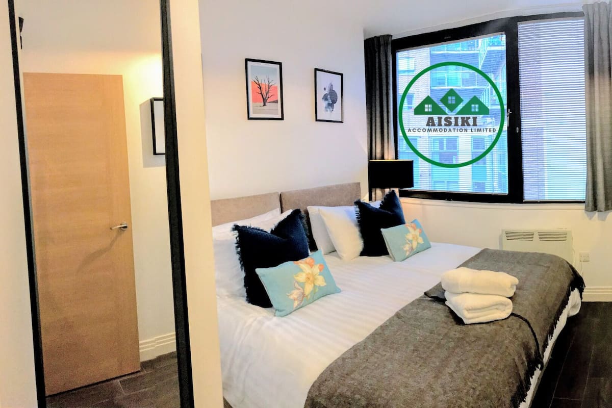 A hotel room with a bed, pillows, a window with blinds, a door, and a wall-mounted air conditioning unit.