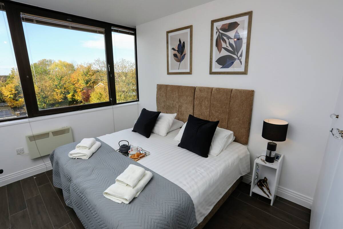A neatly made bed with a grey and white duvet, two black pillows, and a bedside lamp. There are two framed leaf artworks on the wall.